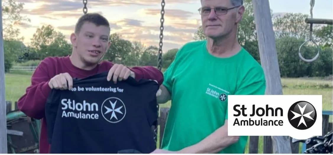 Two men, one younger and one older, stand together outdoors, with the younger man holding up a black shirt with 'St John Ambulance' printed on it, while the older man wears a green shirt also featuring the St John Ambulance logo.